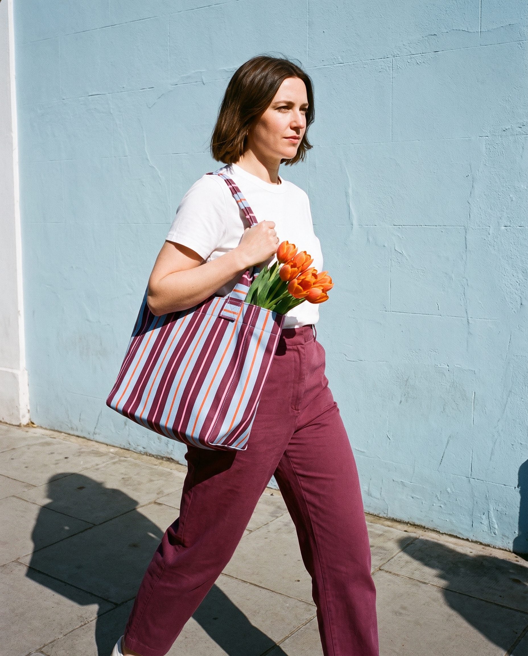A woman walking past a pale blue wall carrying a striped Cassia tote with flowers
